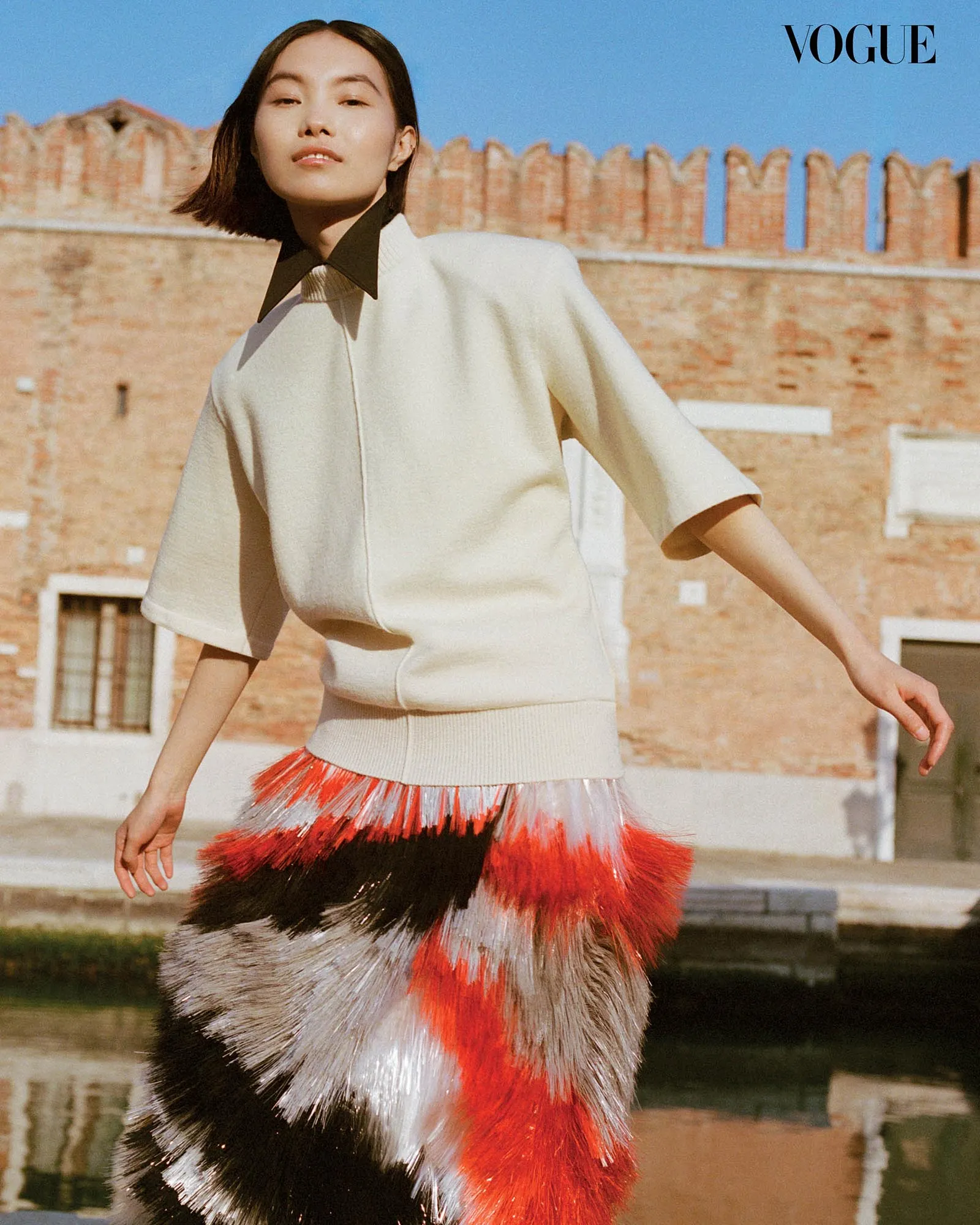 Model wearing a cream sweater and a red‑black fringe skirt outdoors with a brick building in the background; Vogue logo in the corner.