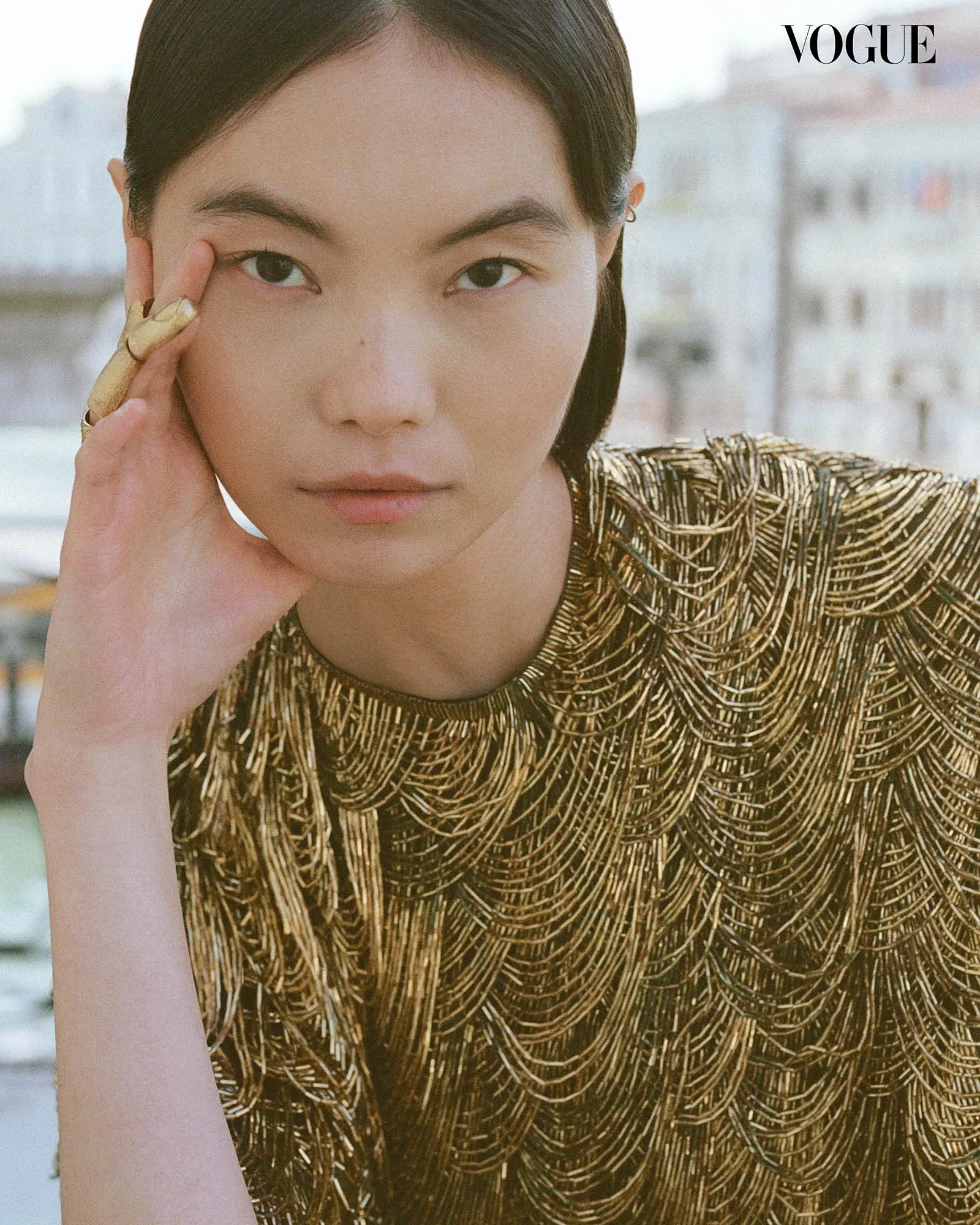 Close-up portrait of a woman resting her cheek on her hand, wearing a gold textured top; Vogue logo in the corner.
