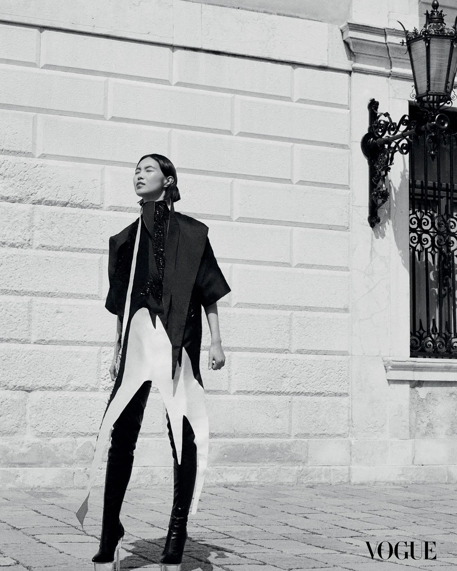 Black-and-white Vogue fashion photo of a model in a bold black-and-white geometric outfit standing on stone steps beside an ornate gate.