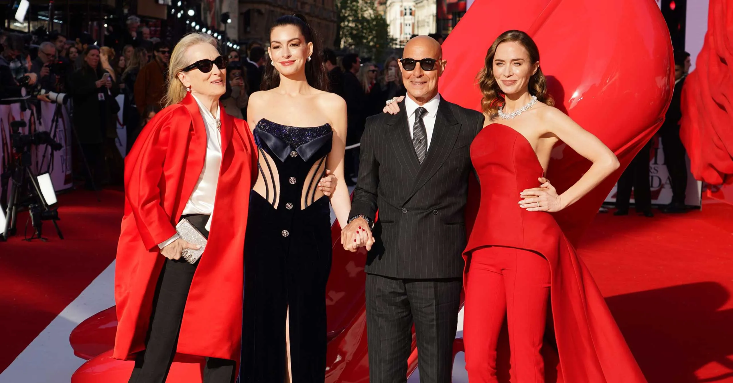 Group of four posing on a red carpet in formal gowns and a suit, photographers in background