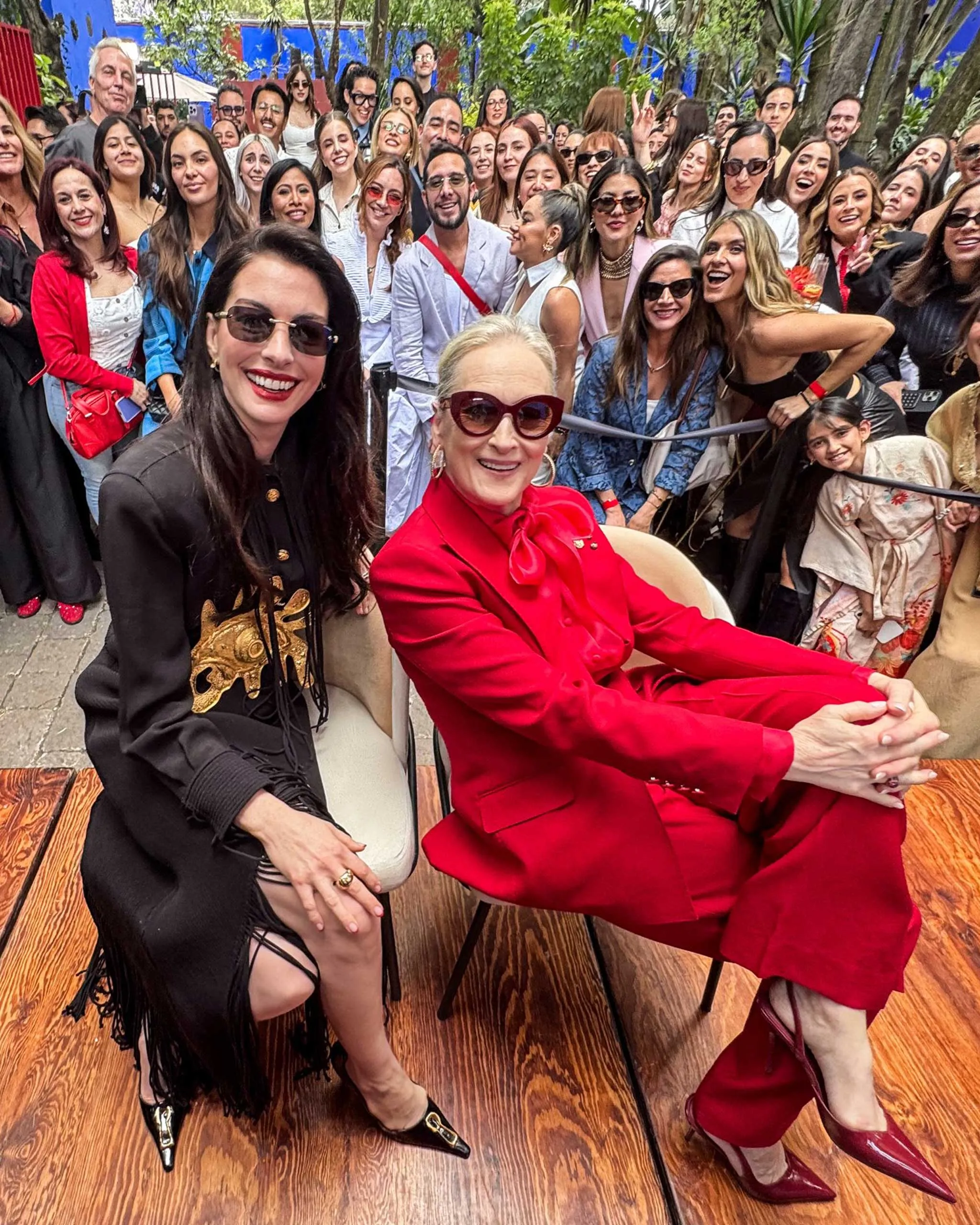 Group selfie at an outdoor event with a crowd behind two women in front wearing bold outfits (red suit and black attire).