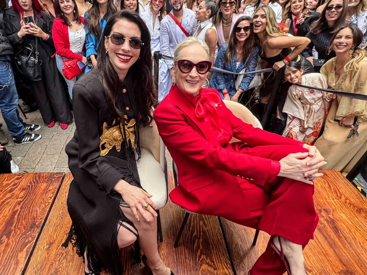 Two women sit at a wooden table in the foreground, smiling; one wears a red suit and sunglasses, the other a black outfit with gold embroidery, as a crowd poses behind them.