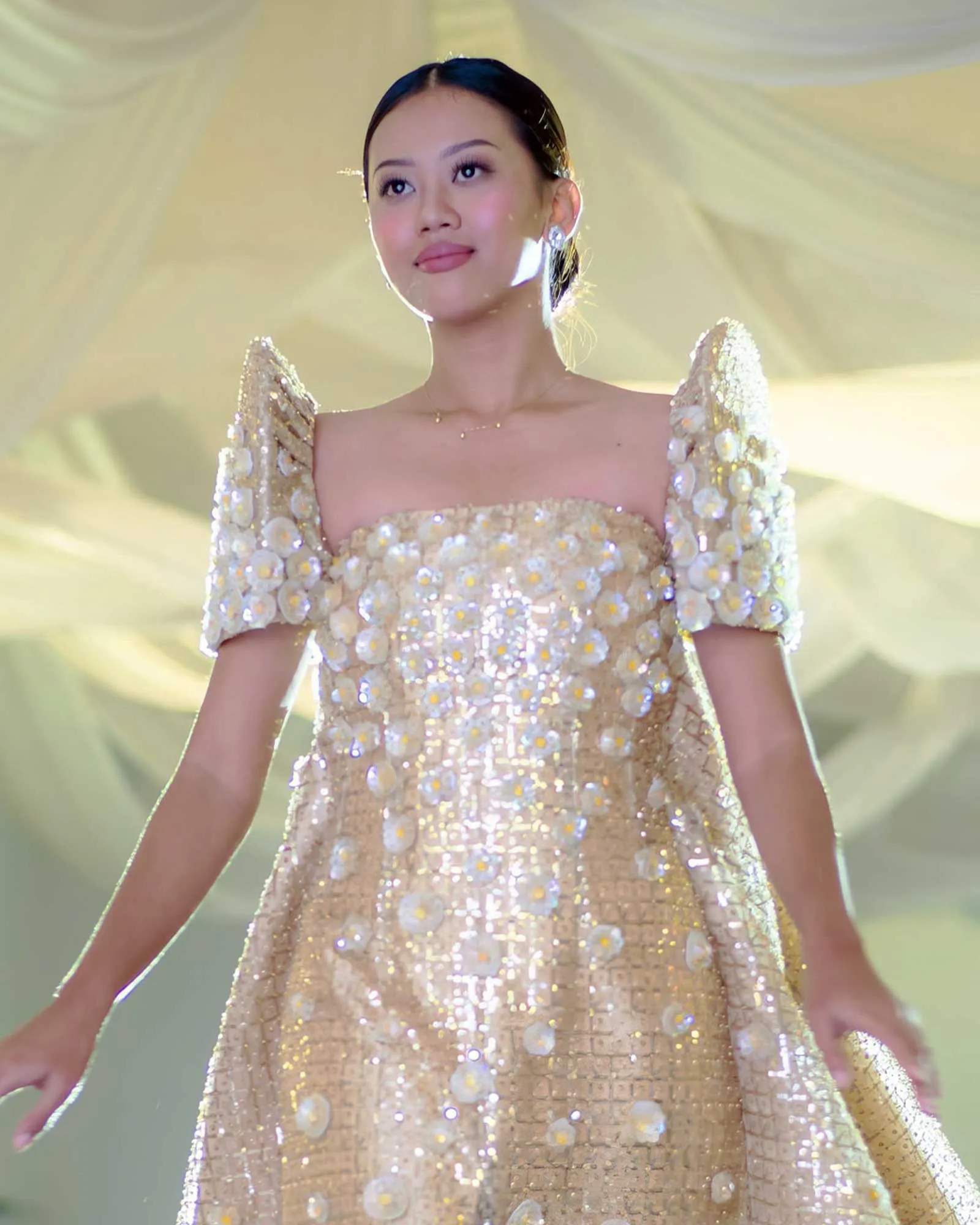 Asian woman in a glittering gold sequin gown on stage, looking upward with soft expression.