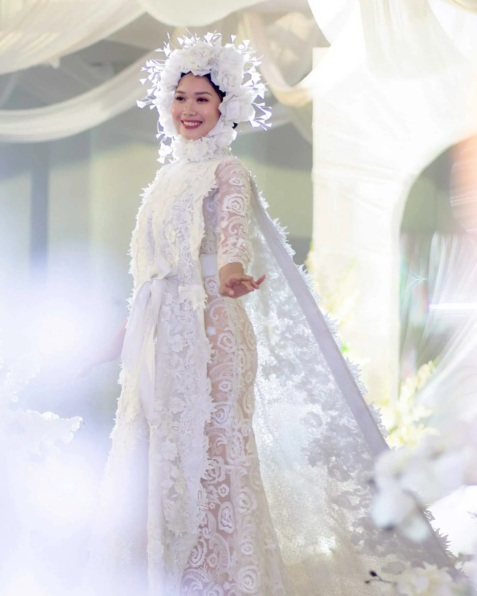 Smiling person in a white lace wedding dress with a large floral headpiece.