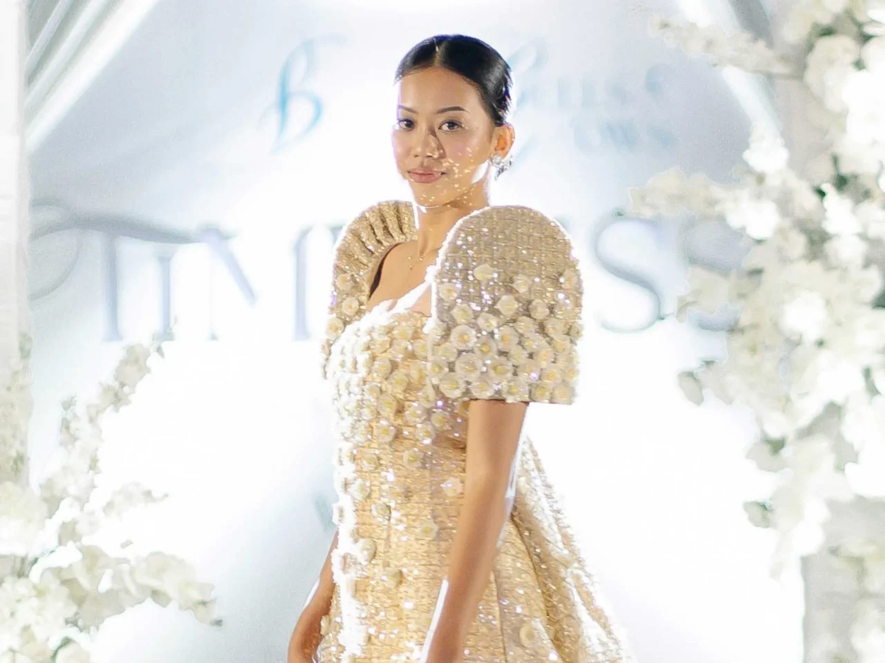woman wearing a gold sequin outfit with floral backdrop, posing at a bright event
