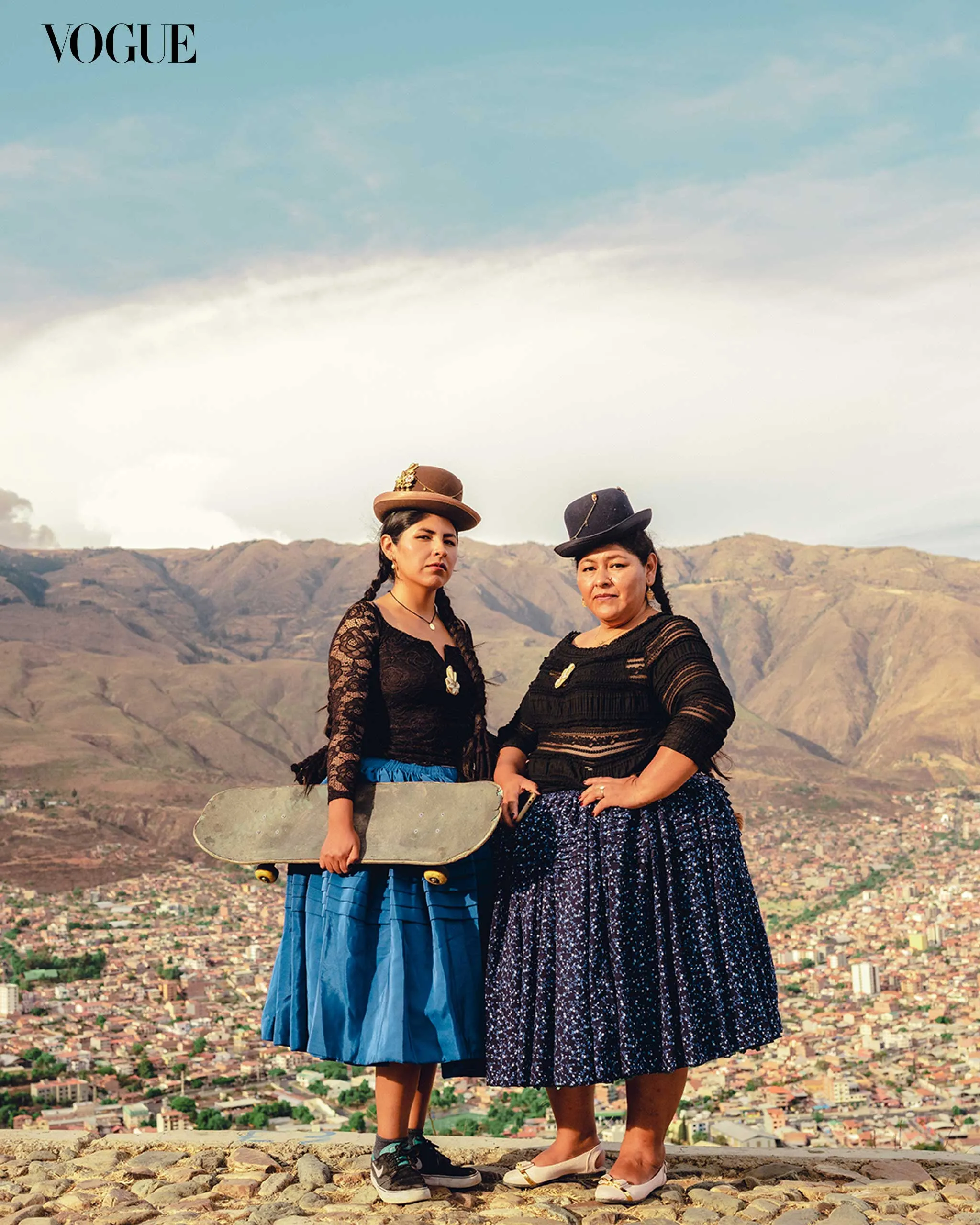 Two women in traditional clothing stand on a rocky overlook, with a city and mountains in the distance; Vogue logo in the top-left corner.