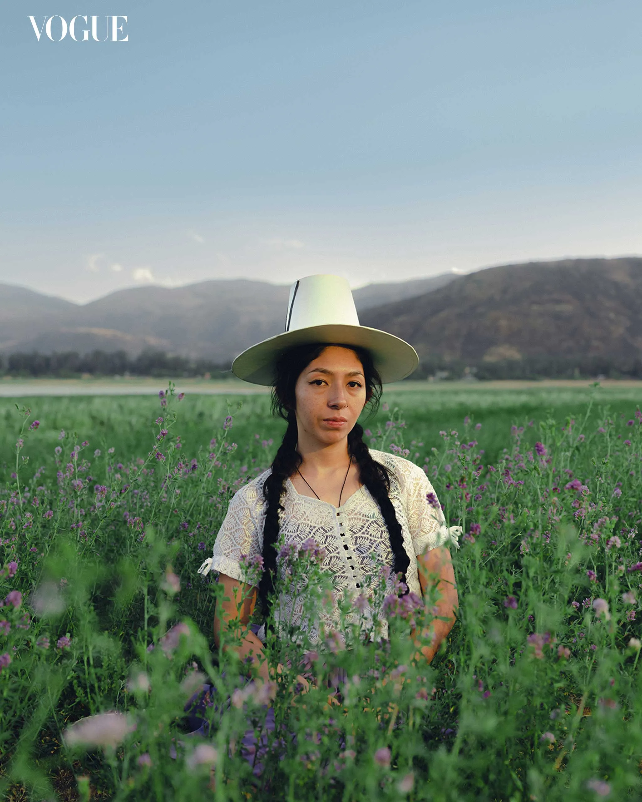 Woman with braided hair and a wide-brim hat stands in a lavender field, wearing a lace blouse, mountains in the distance, Vogue logo upper-left.