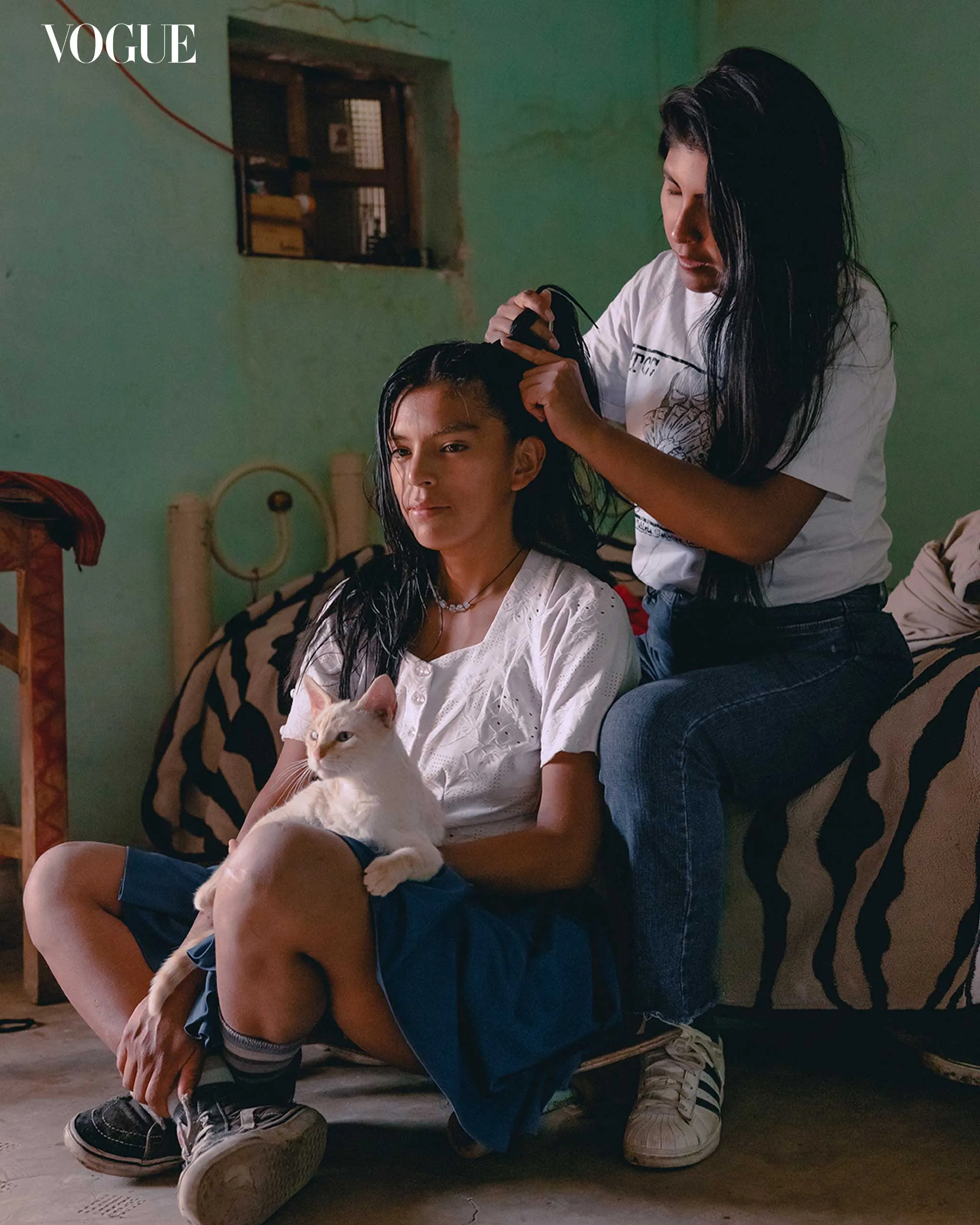 Two young women in a green room; one braids the other's hair while a white cat sits on her lap.