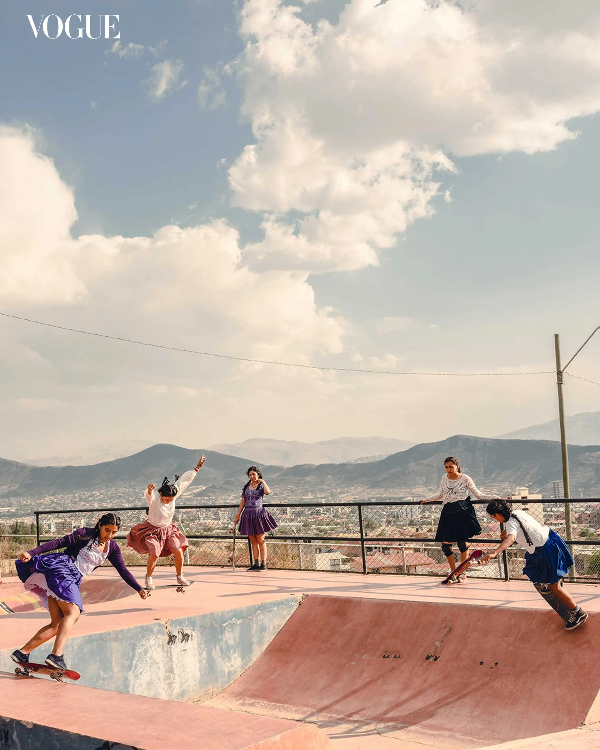 Group of young women skateboarding and posing on a pink concrete ramp at a sunny outdoor skatepark, mountains in the background, Vogue watermark in the corner.