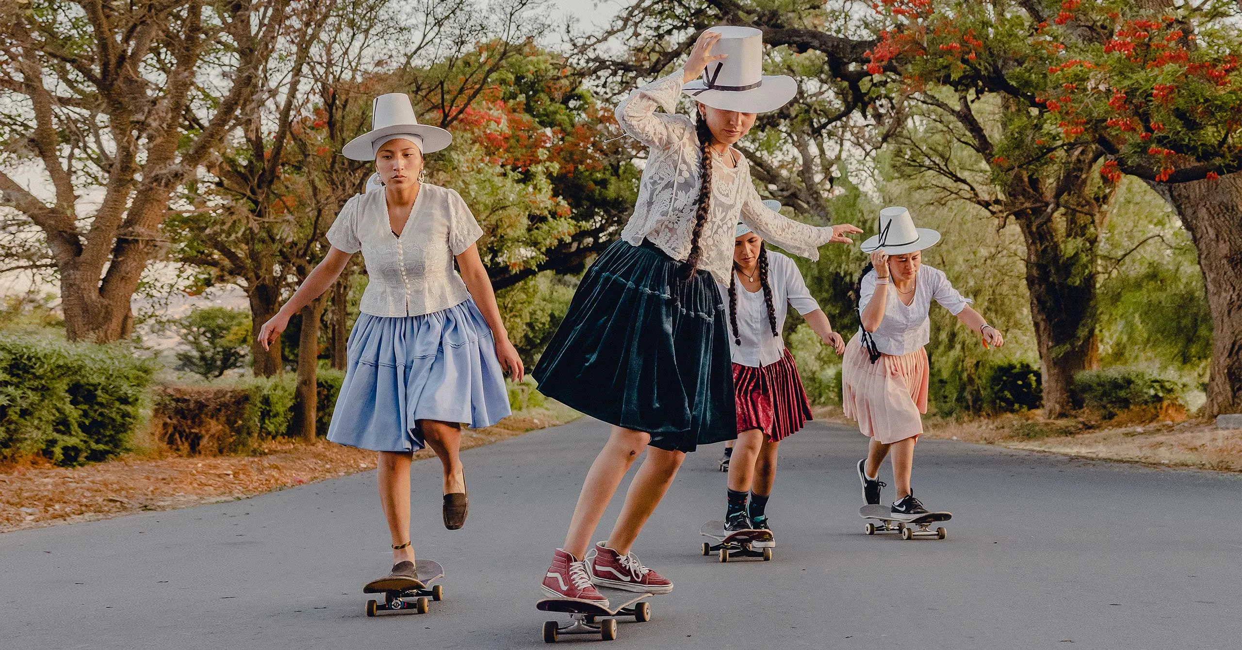 Group of young women skateboarding together on a tree-lined road in vintage white tops, skirts, and matching wide-brim hats.