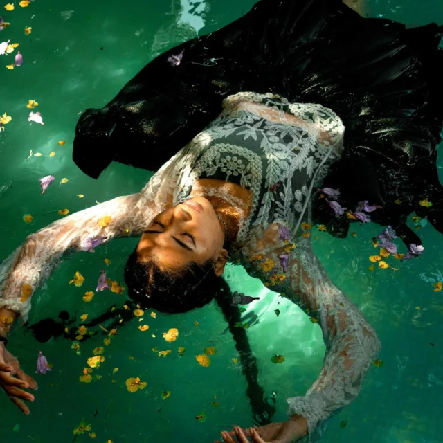 Woman in a white lace dress floats on green water, surrounded by scattered flower petals.
