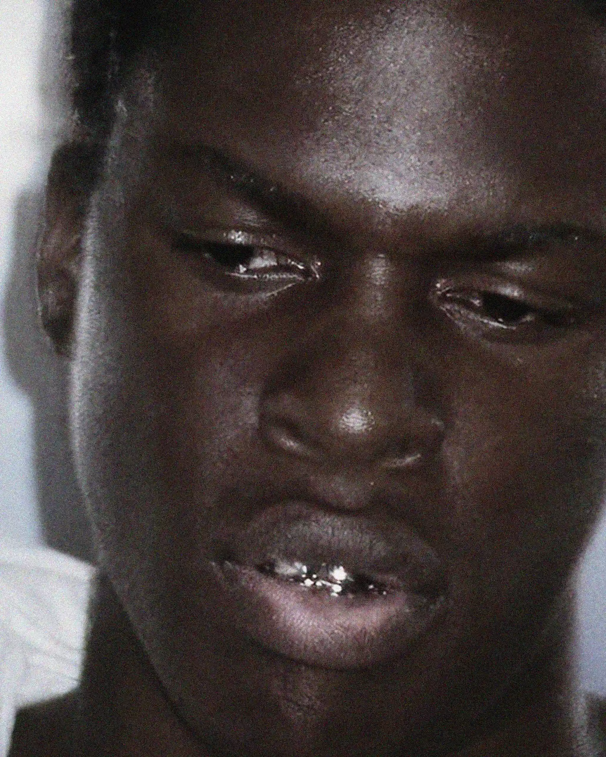 Close-up of a young man's face, sweating, looking downward, with a metallic grill on his teeth