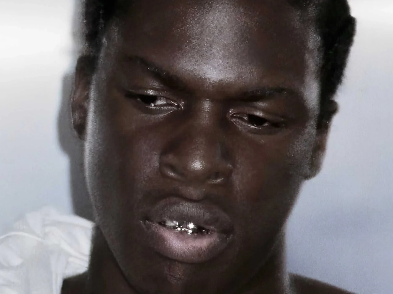 Close-up of a young man with a dark complexion, looking downward with a grimace and a metal grill on his teeth.