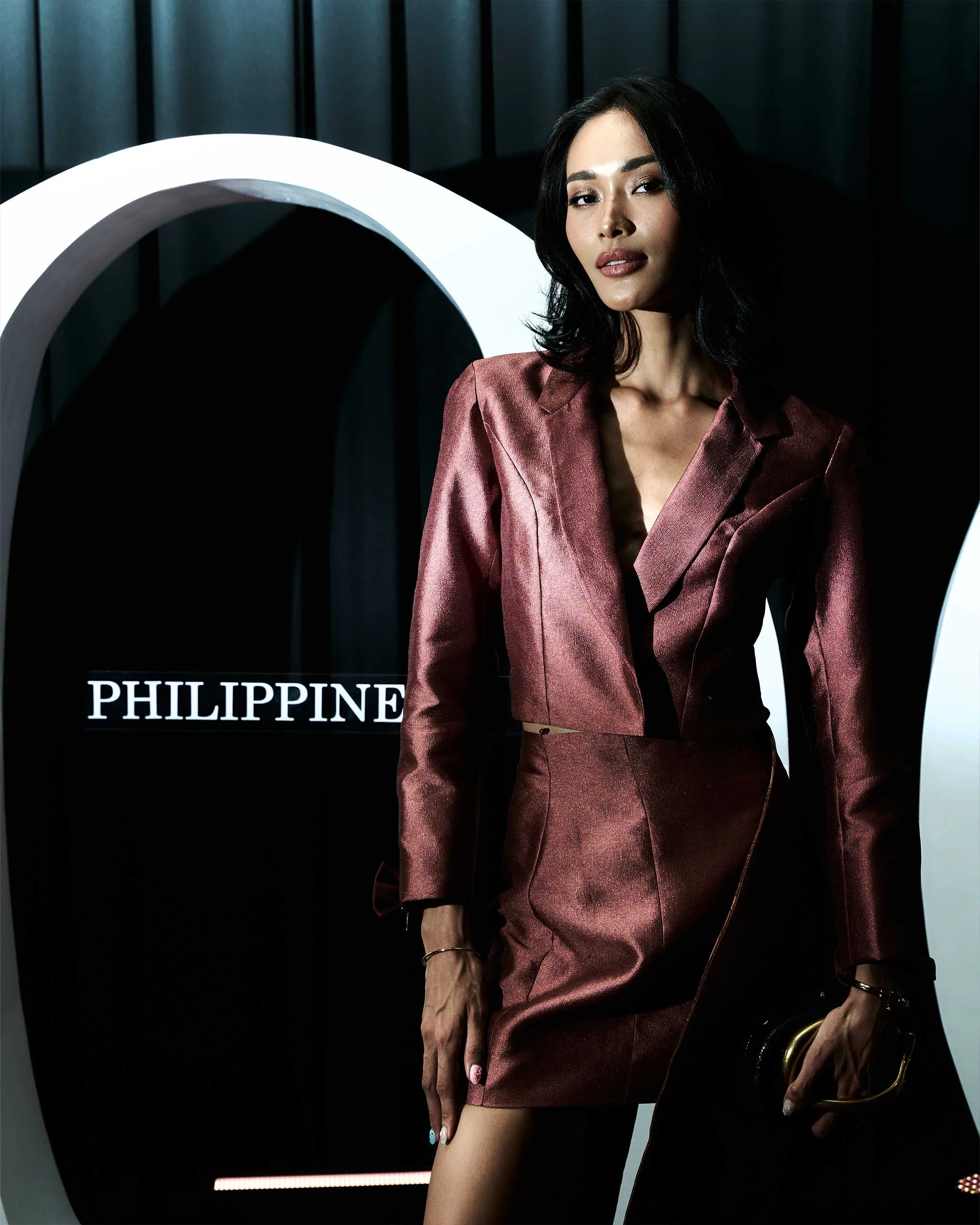 Model in a metallic pink blazer and skirt stands against a dark backdrop with a large white curved prop behind her, during a fashion shoot.