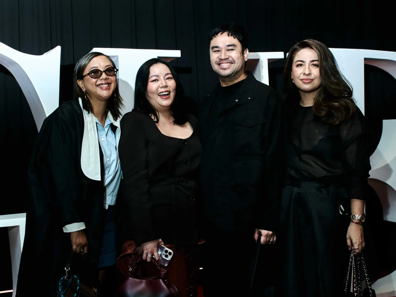 Six stylish adults stand together at an event, wearing bold black, gray, and metallic outfits against a dark backdrop with a large white logo behind them.
