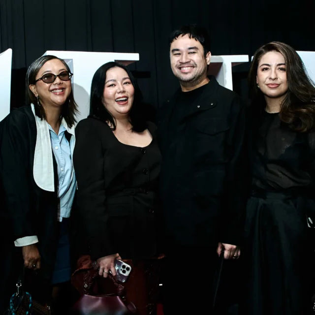 Four adults in black outfits posing together at an event, smiling with large white letters in the background behind them.