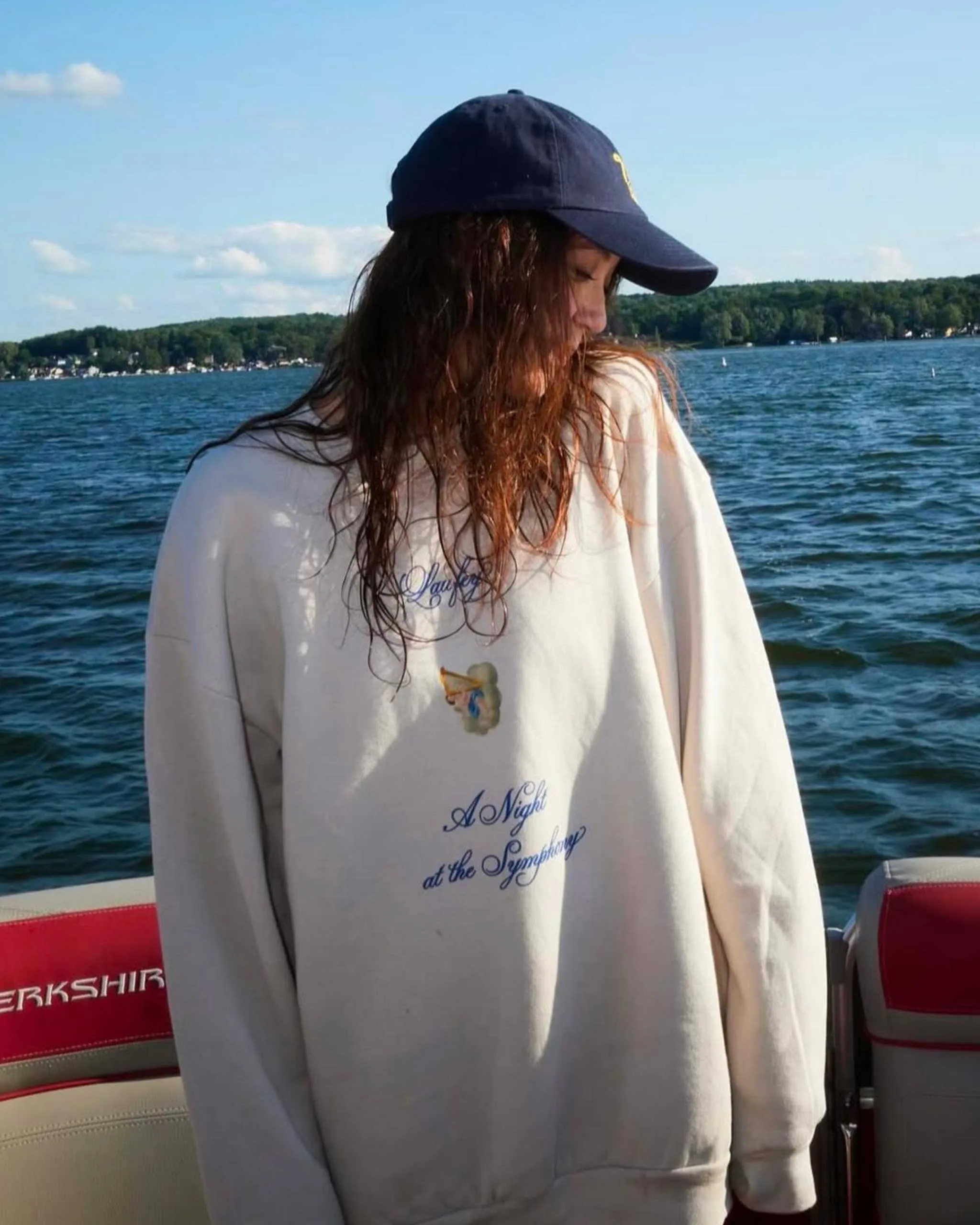 Person with long wet hair wearing a navy baseball cap and oversized light sweatshirt, standing on a boat with a lake and shoreline in the background.