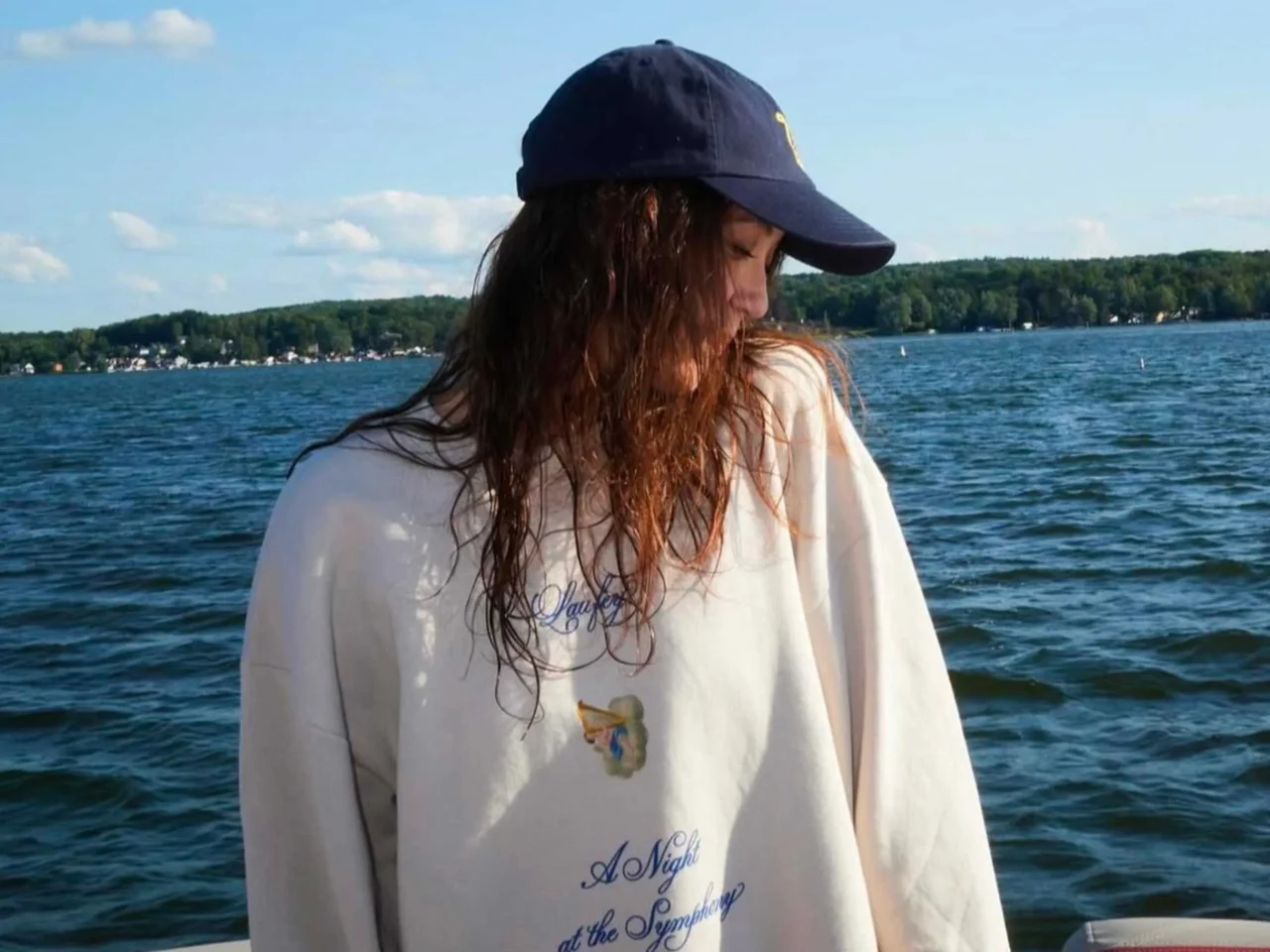 Person in a navy cap and white shirt with long wet hair stands in a lake, tree-lined shore in the background.