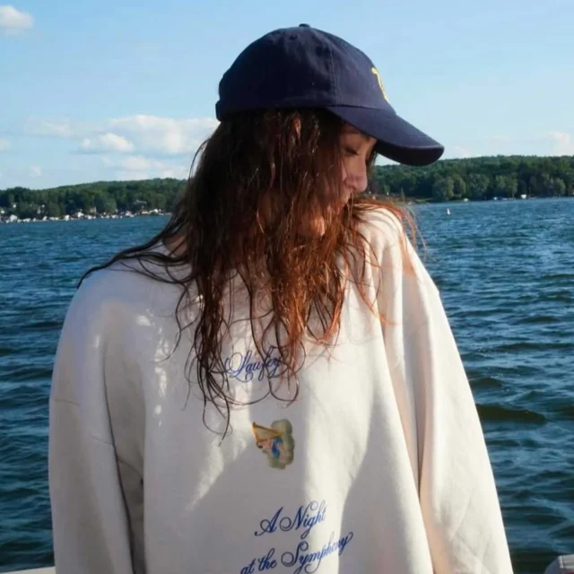 Person with a dark cap and long hair on a boat, looking down, blue lake and tree-lined shore in the background.