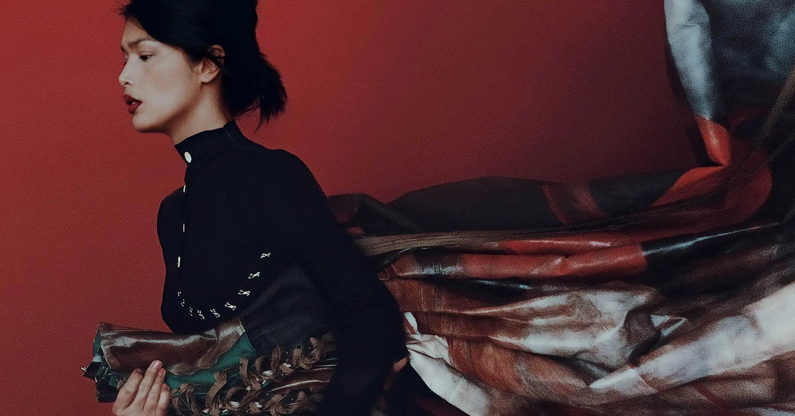 Side profile of a person with dark hair in a black outfit against a solid red backdrop, holding a multicolored patterned scarf that billows to the right.