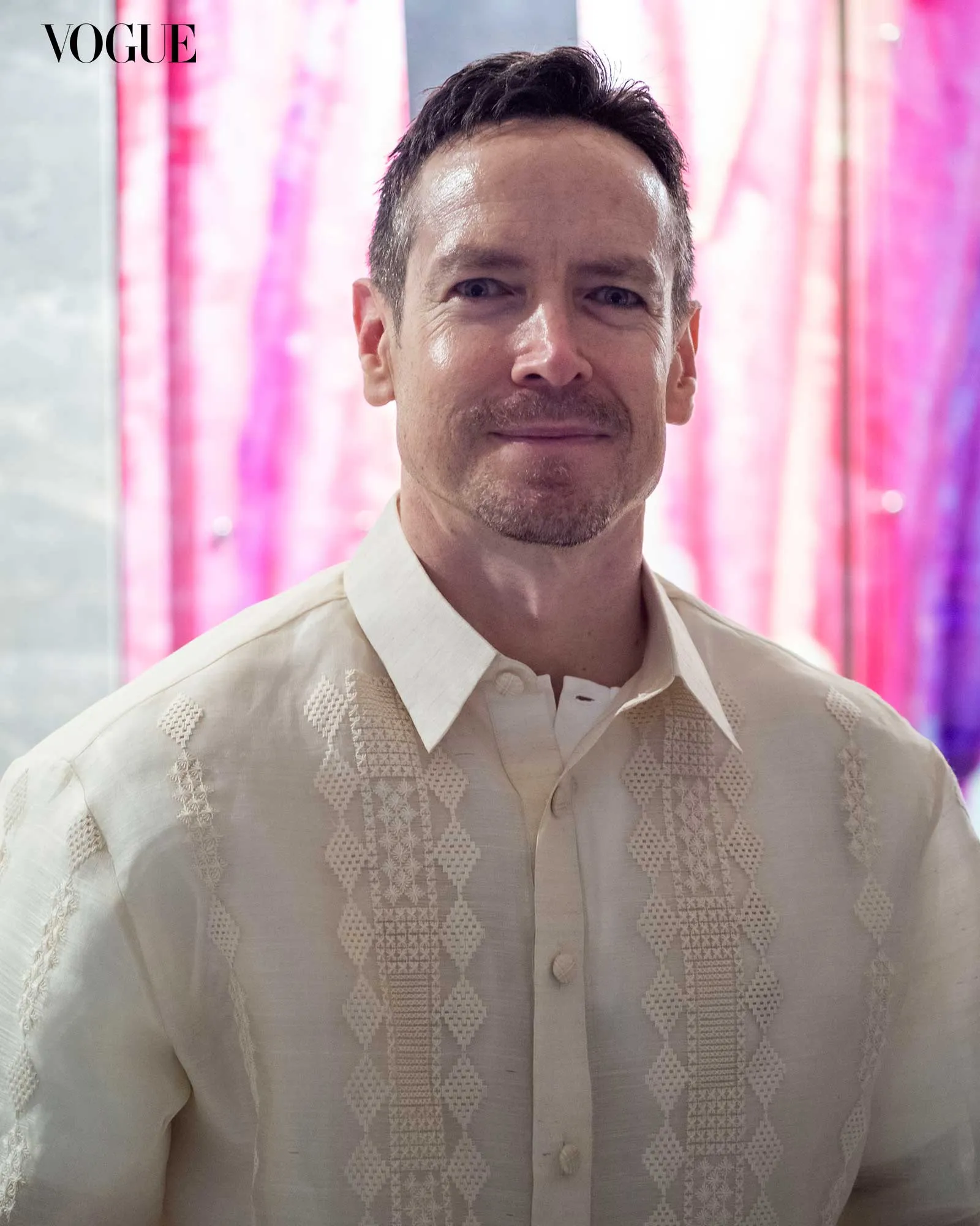 Portrait of a man in a cream embroidered shirt, smiling lightly, with pink light backdrop and a Vogue logo in the corner.