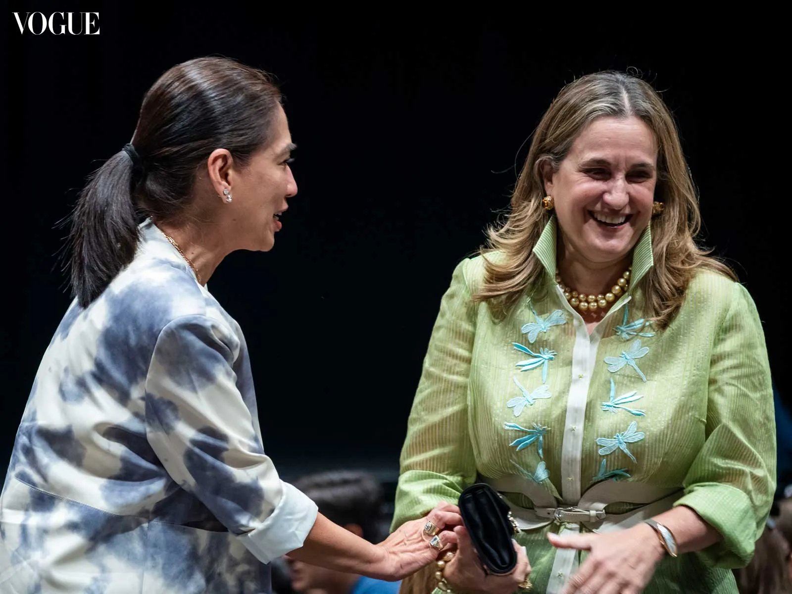 Vogue logo in upper left; two women smiling and shaking hands on stage, left in a tie-dye shirt and right in a light-green embroidered blouse with dragonfly designs and a pearl necklace.
