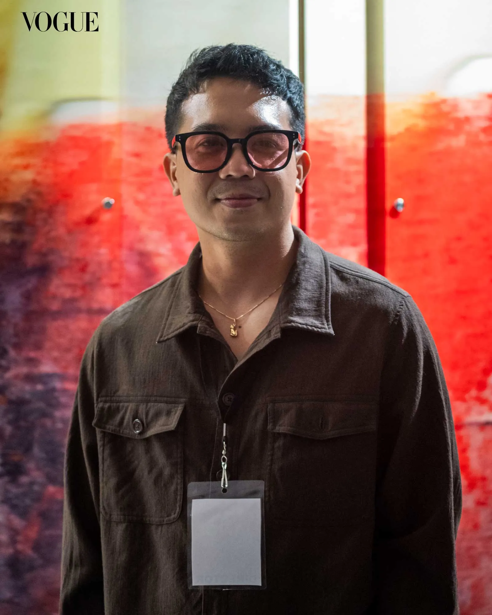 Portrait of a man in a dark shirt wearing glasses, with a lanyard and badge, in front of a red-orange gradient backdrop (Vogue logo visible).