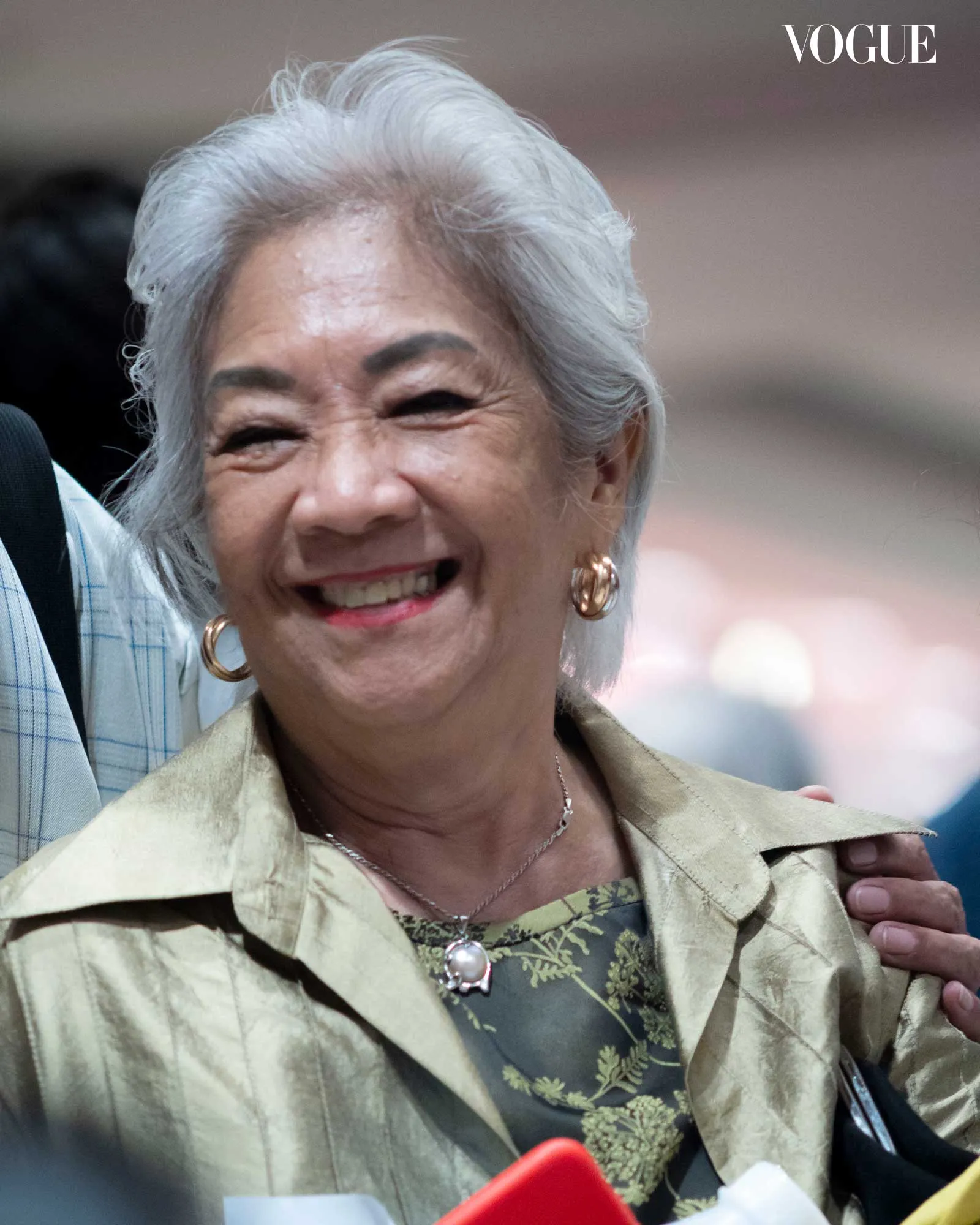Smiling elderly woman with short gray hair and gold hoop earrings, wearing a beige jacket over a floral dress; Vogue logo in the top-right corner.