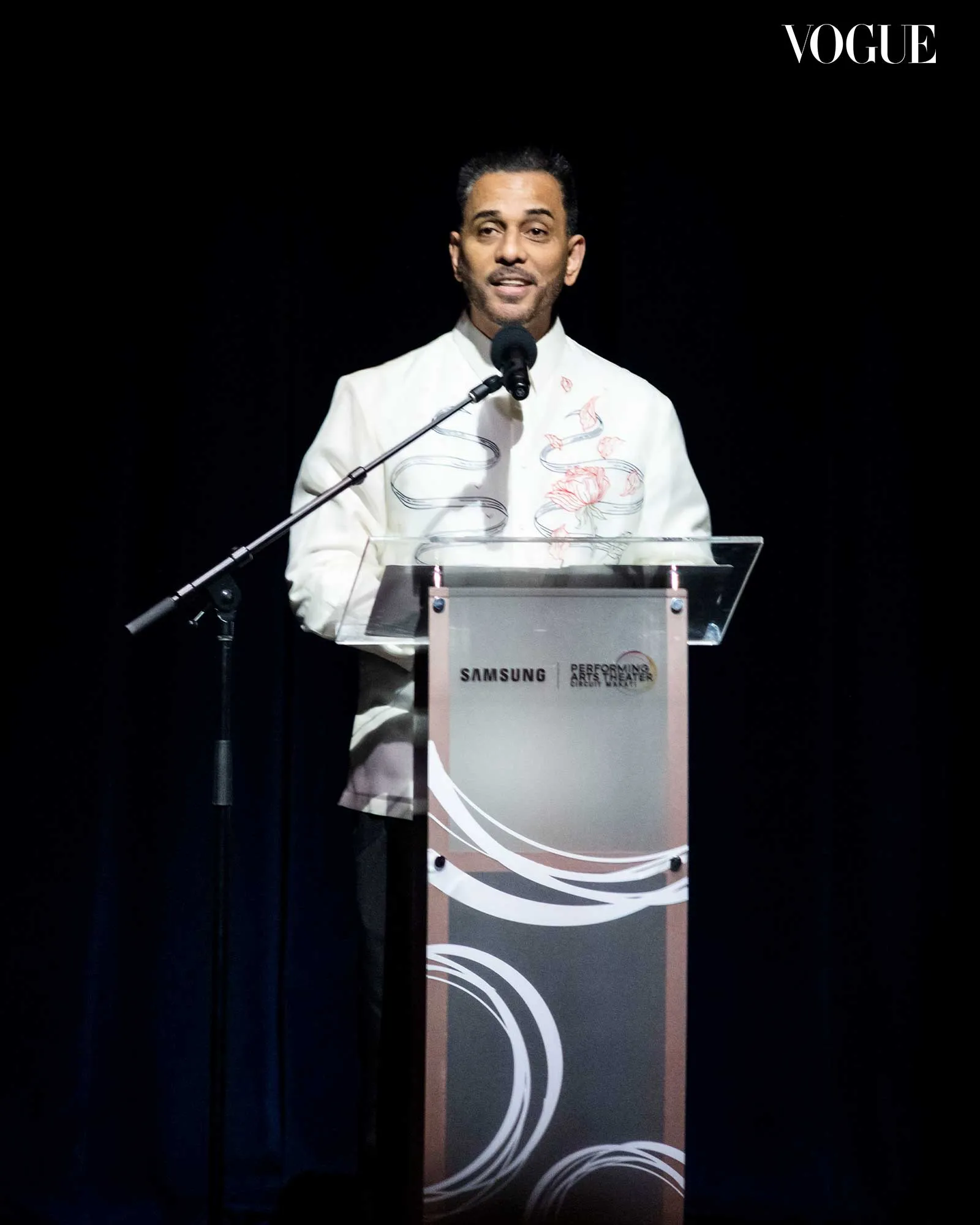 Man in a white jacket speaks at a podium on a dark stage; Vogue logo in the top-right corner and Samsung branding on the lectern.
