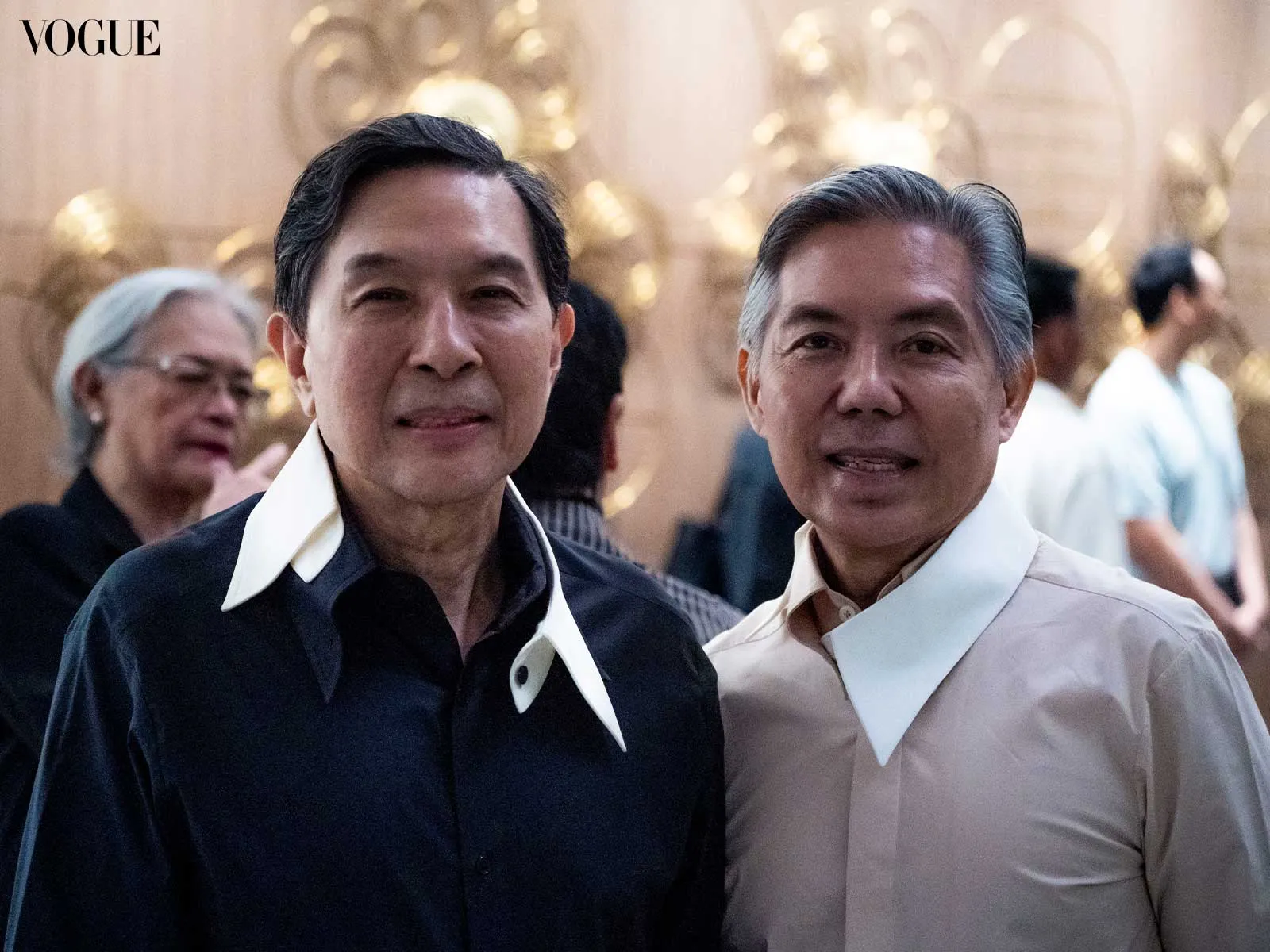 Two men pose for a photo at a Vogue event, wearing dark and light collared shirts with distinctive white neck collars