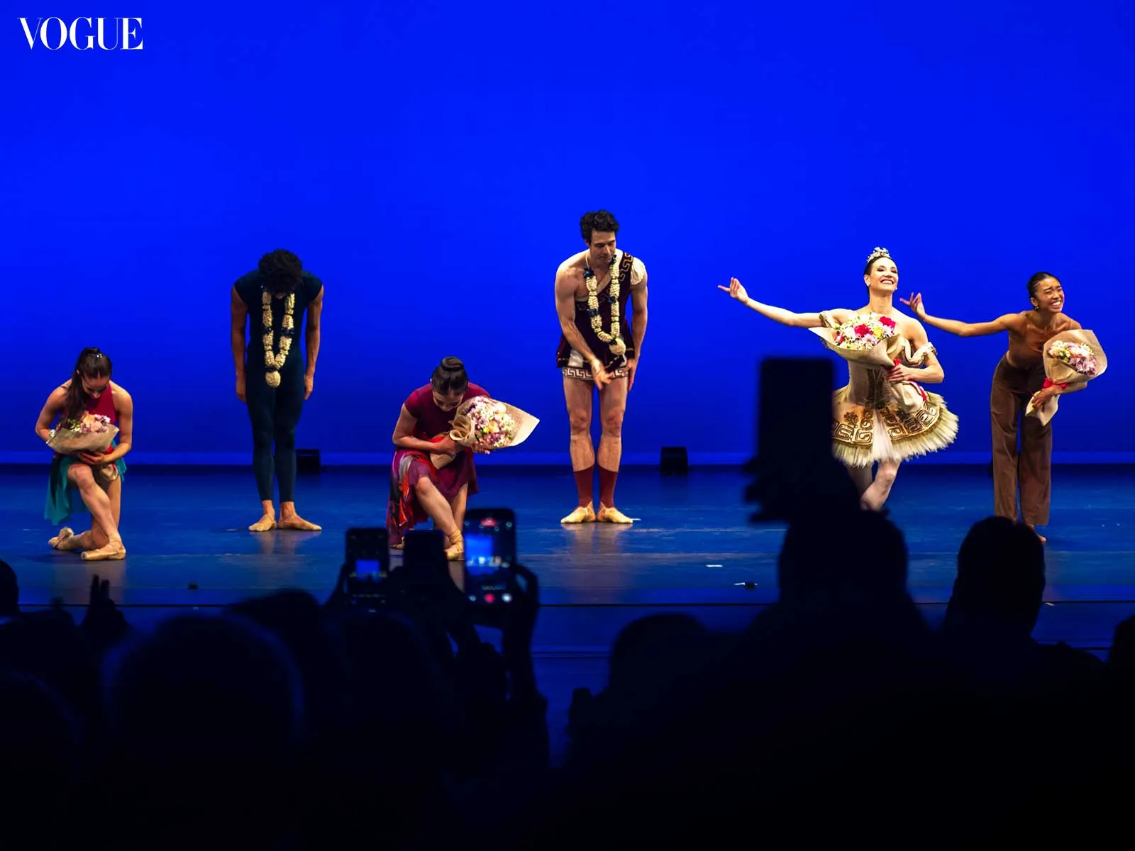 Six dancers bow on a bright blue stage, holding floral bouquets; Vogue logo appears in the top-left corner.