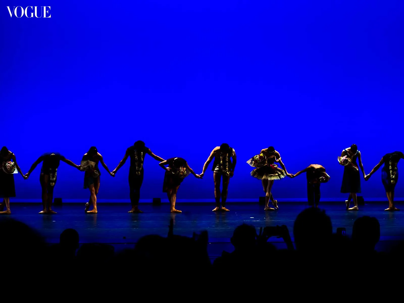 Silhouetted dancers on a blue stage hold hands in a line, bows at the end of a performance; Vogue logo in top-left corner.