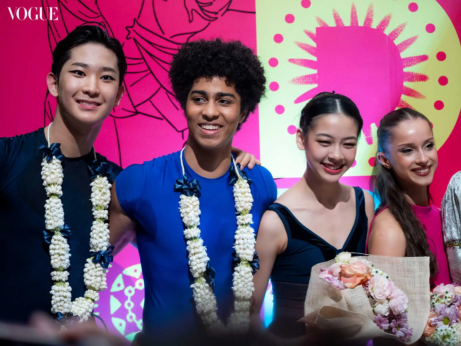 Four models with flower garlands pose on a bright pink and lime backdrop, Vogue logo visible in the corner.