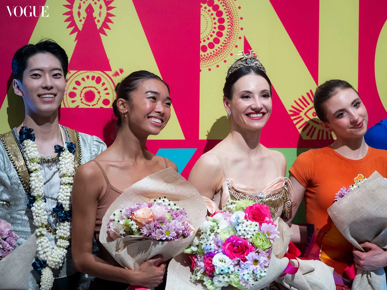 Four models pose with bouquets at a Vogue event, smiling against a bright pink and lime backdrop with the Vogue logo visible.