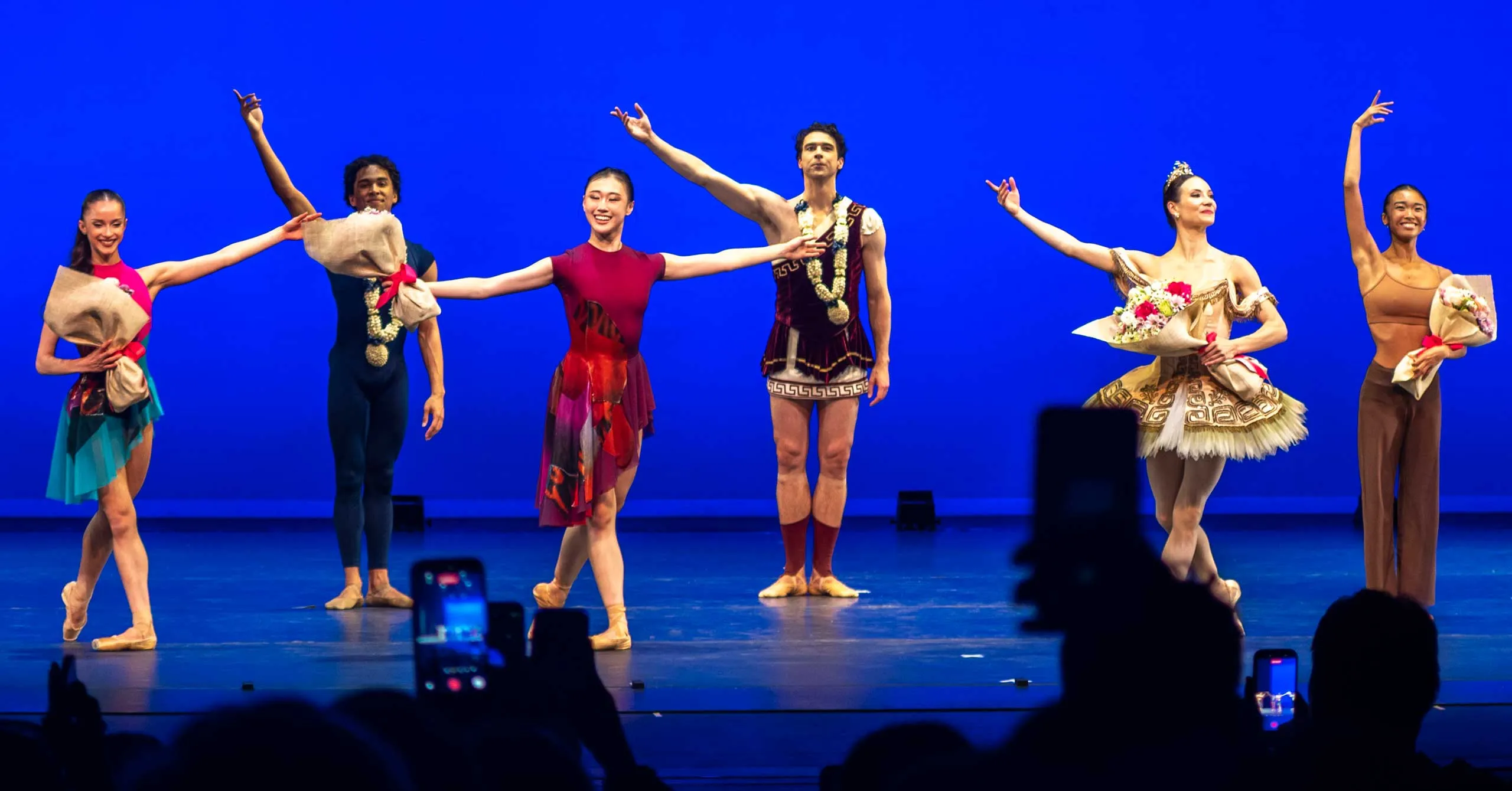 Six ballet dancers stand in a line on a blue stage, each holding bouquets wrapped in brown paper, after a performance. Audience members with phones are visible in the foreground.