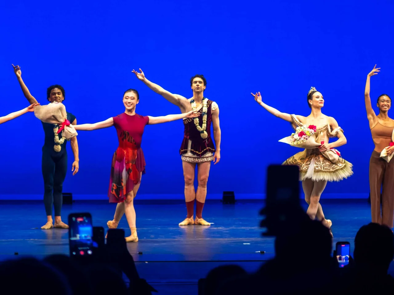 Six ballet dancers stand in a line on a blue stage, each holding bouquets wrapped in brown paper, after a performance. Audience members with phones are visible in the foreground.