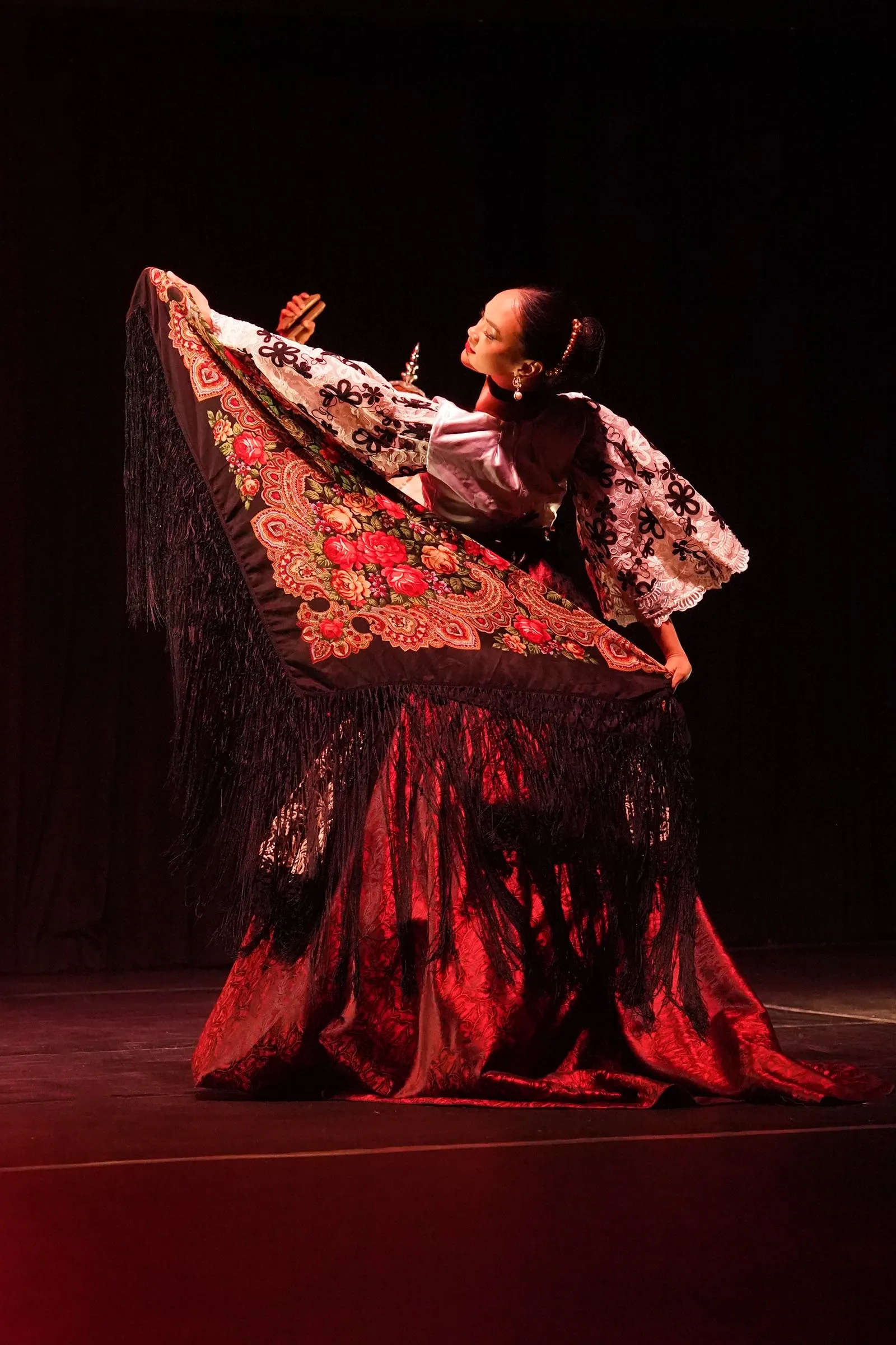 Flamenco dancer in a red and black embroidered dress with a large shawl, performing on stage under red lighting.