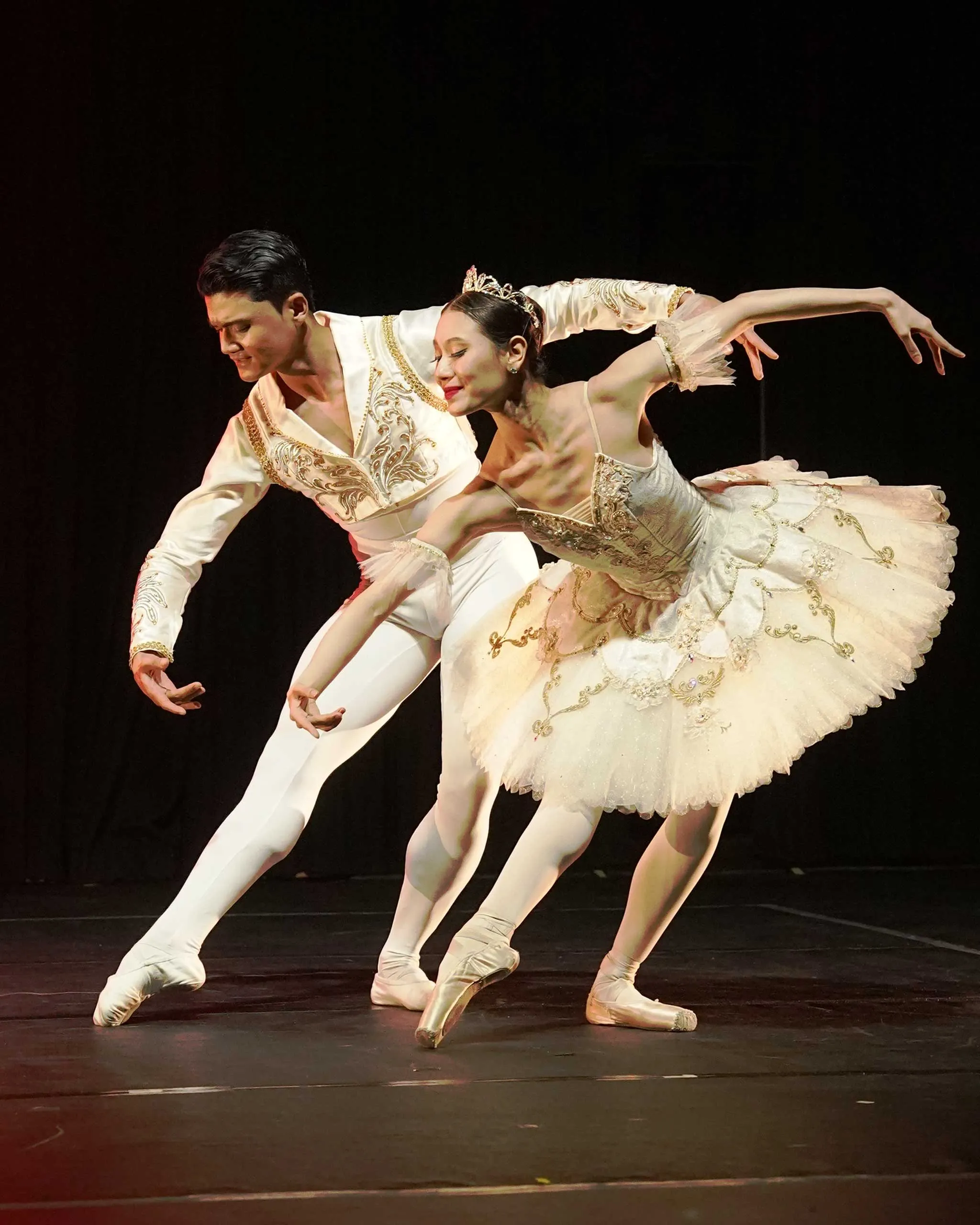 Two ballet dancers in white costumes performing a synchronized pose on stage, graceful arm extensions and pointe shoes.