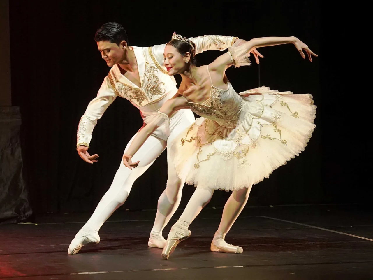 Two ballet dancers in white and gold costumes perform a duet on a dark stage, arms extended gracefully.