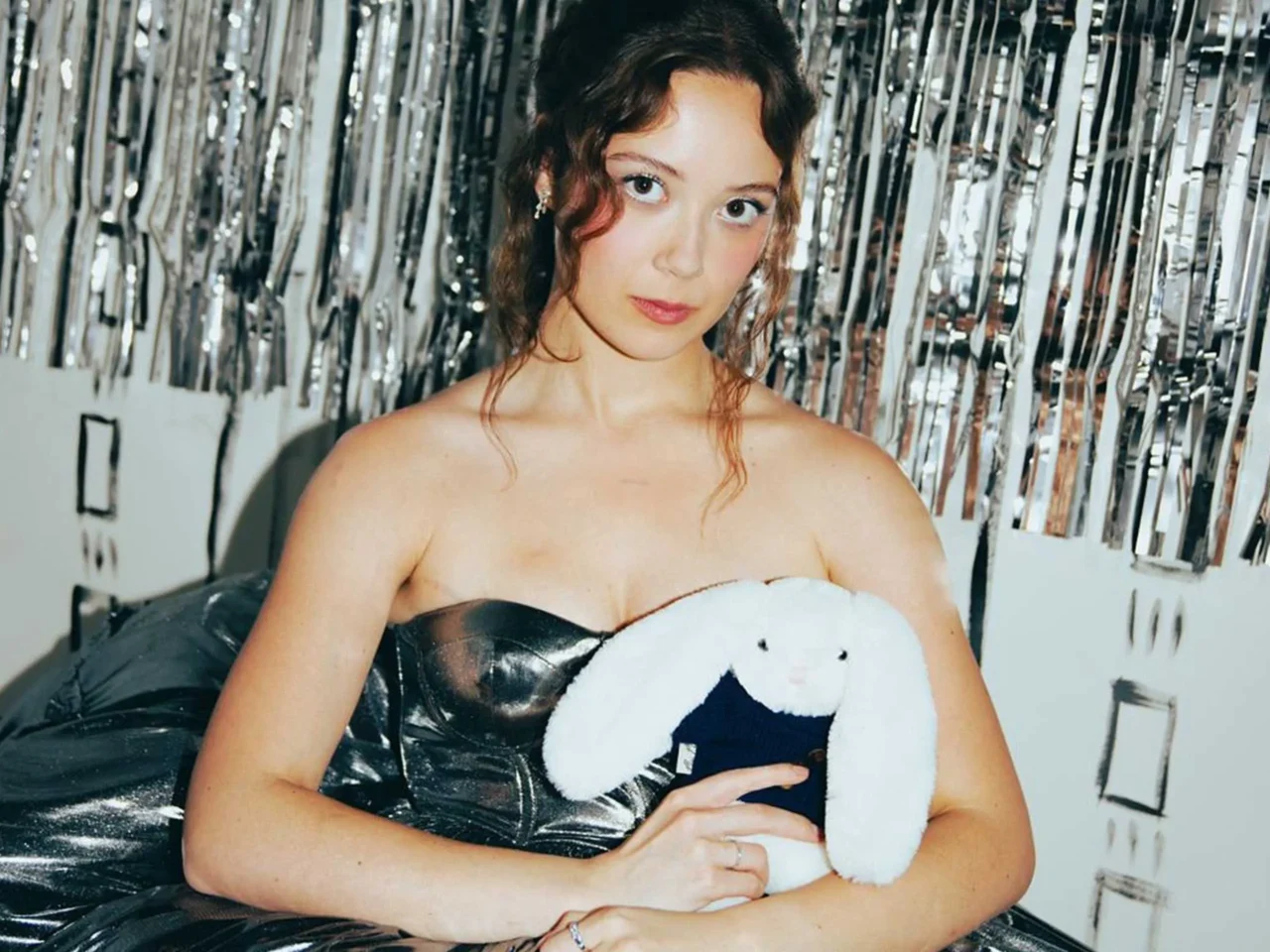 Portrait of a young woman with brown hair posing against a shiny silver tinsel backdrop, looking at the camera with a soft expression.