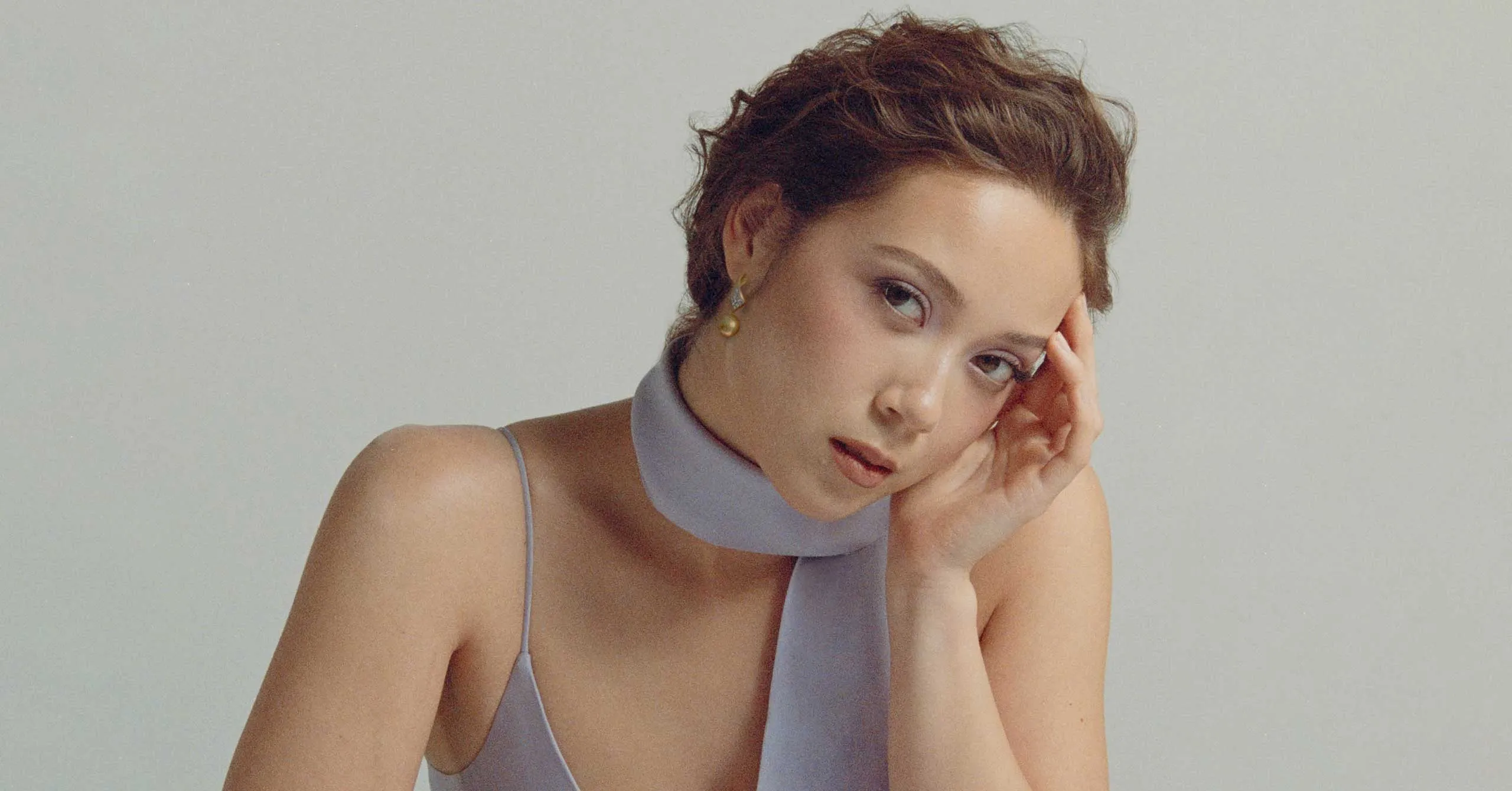 Young woman with short hair rests her head on a hand, wearing a pale blue halter top, against a neutral background.