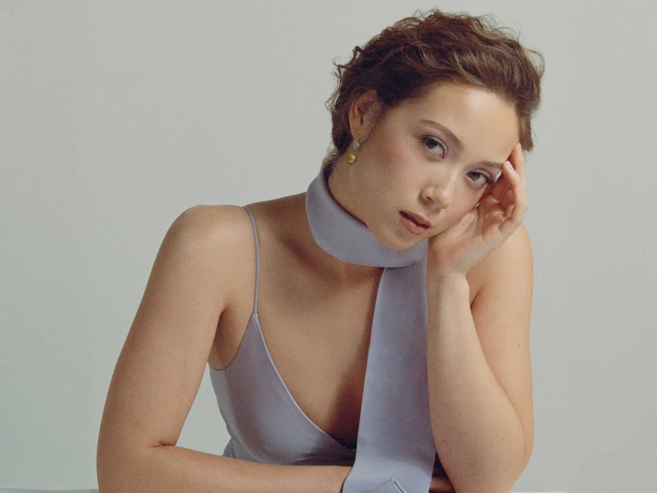 Young woman with short hair rests her head on a hand, wearing a pale blue halter top, against a neutral background.