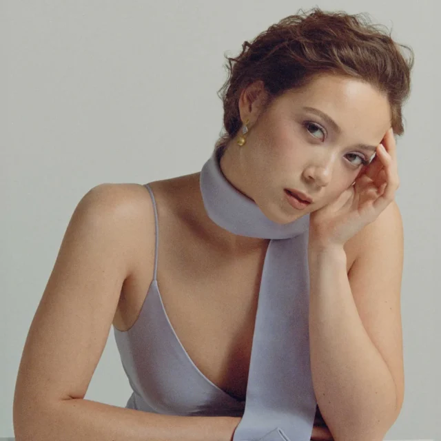 Close-up portrait of a woman with short hair, wearing a light gray halter dress, resting her head on her hand.