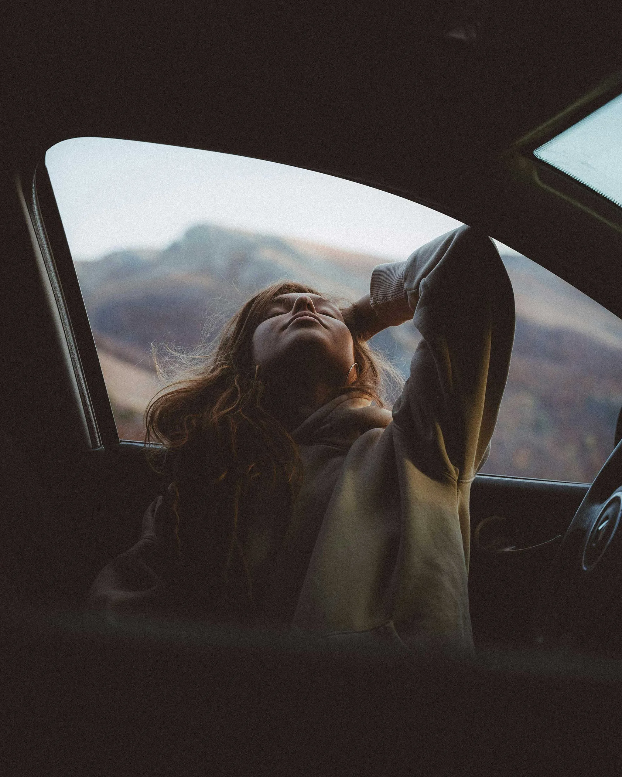 Person leaning back with arms raised through an open car window, mountains in the distance outside.
