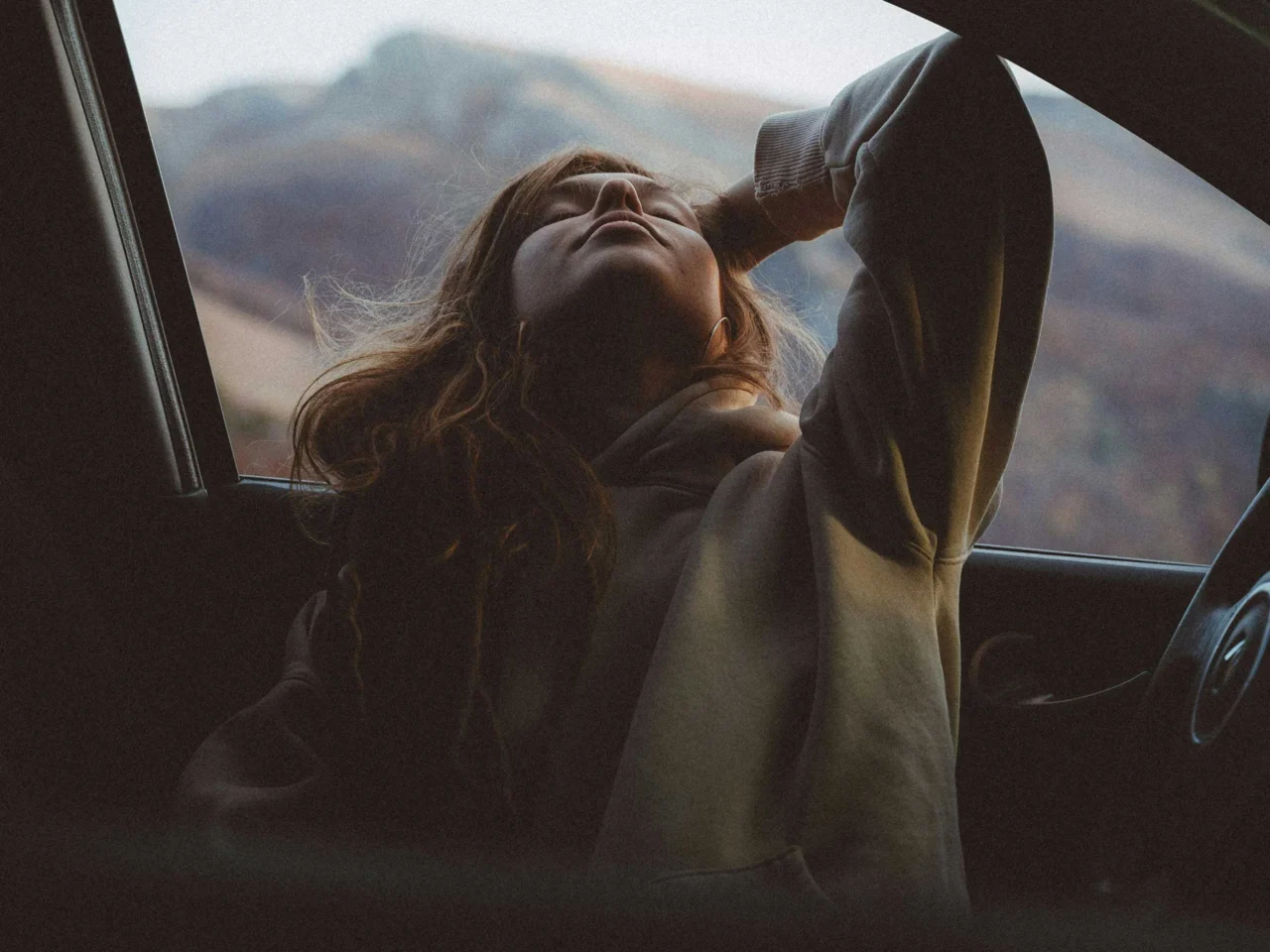 Person leaning out of a car window, face tilted up, with mountains in the background.