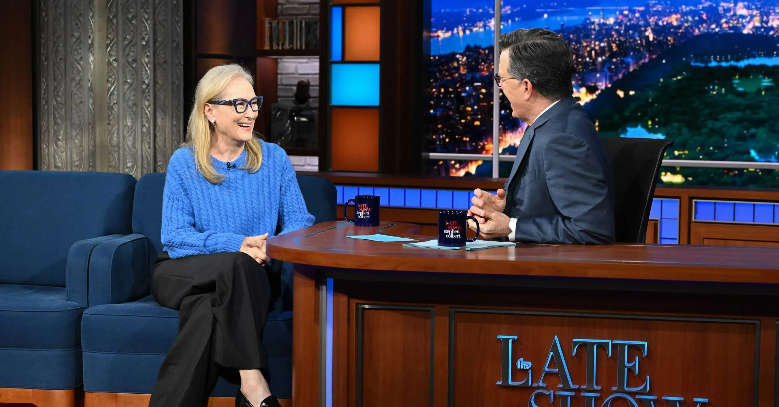 Host and guest smile and chat on The Late Show set, seated on a blue sofa with mugs on a wooden desk and a nighttime city backdrop.