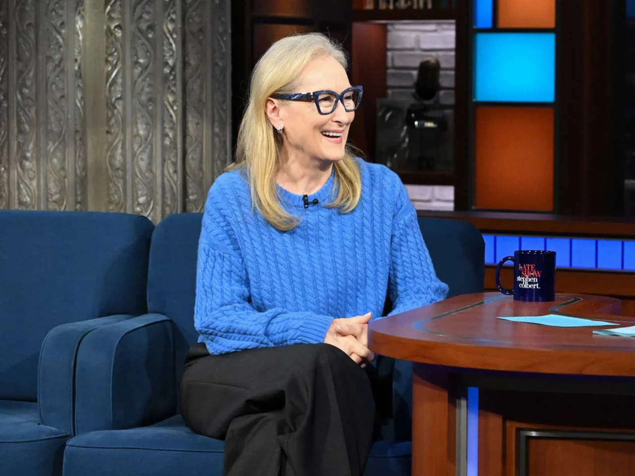 Host and guest smile and chat on The Late Show set, seated on a blue sofa with mugs on a wooden desk and a nighttime city backdrop.