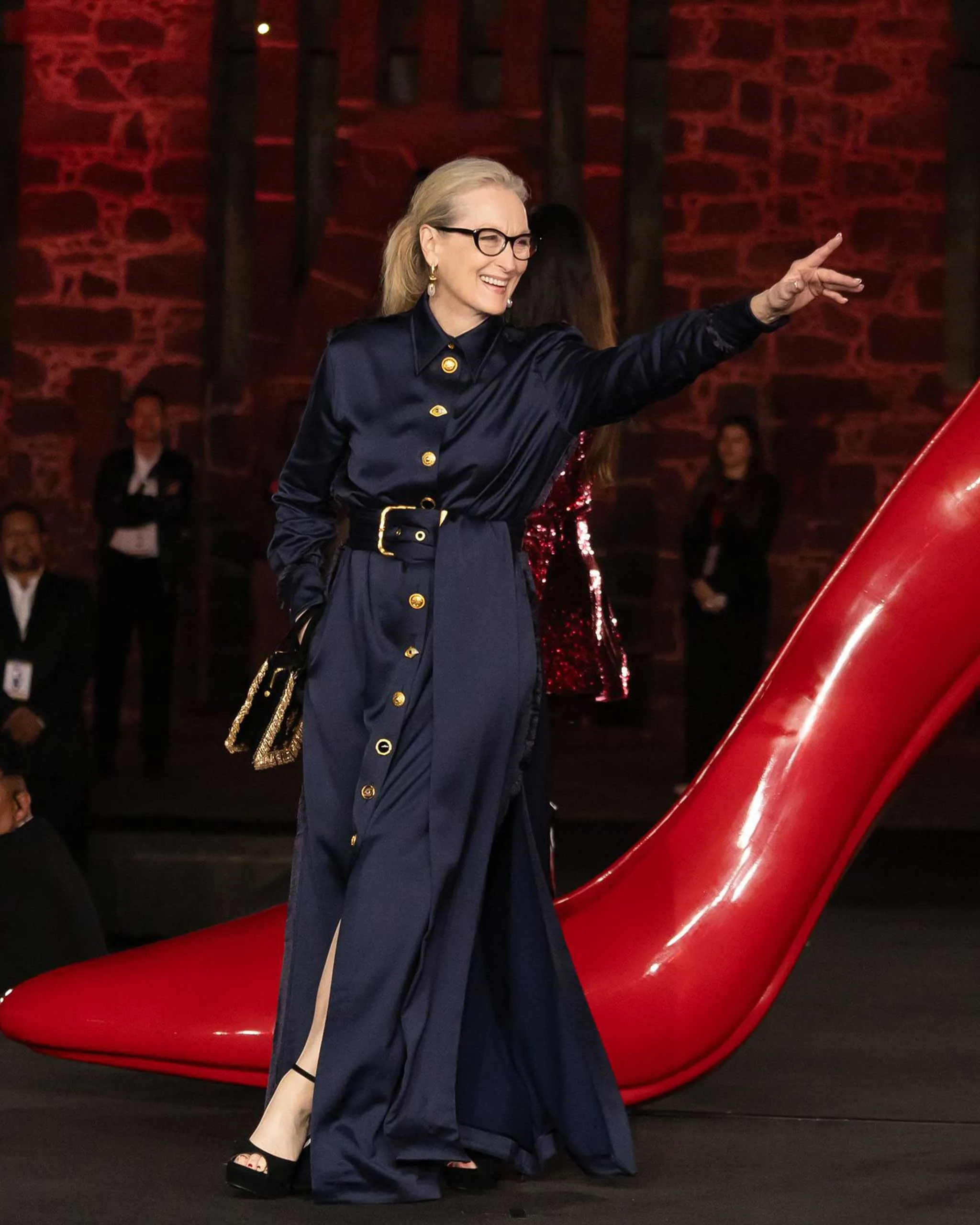Smiling woman in a navy satin dress with gold buttons and a belt walks a runway, pointing to the right with glasses on.