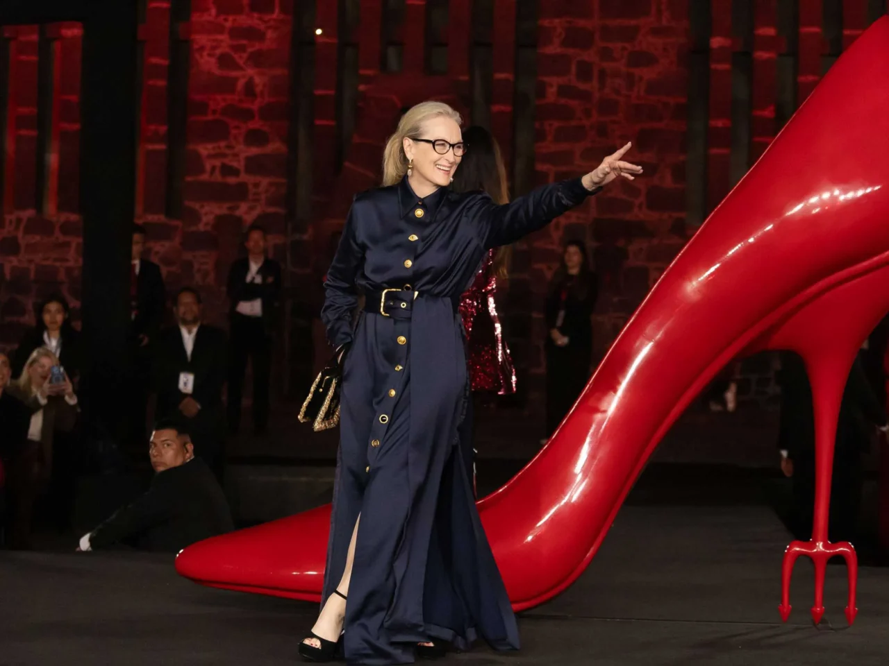 Woman in a navy belted dress with gold buttons walks beside a gigantic red high-heeled sculpture at a fashion show, smiling at the crowd.