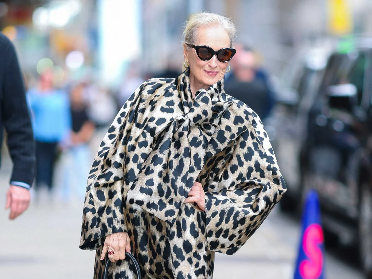 Older woman in a bold leopard-print coat and dark sunglasses, smiling as she strolls through a city street.